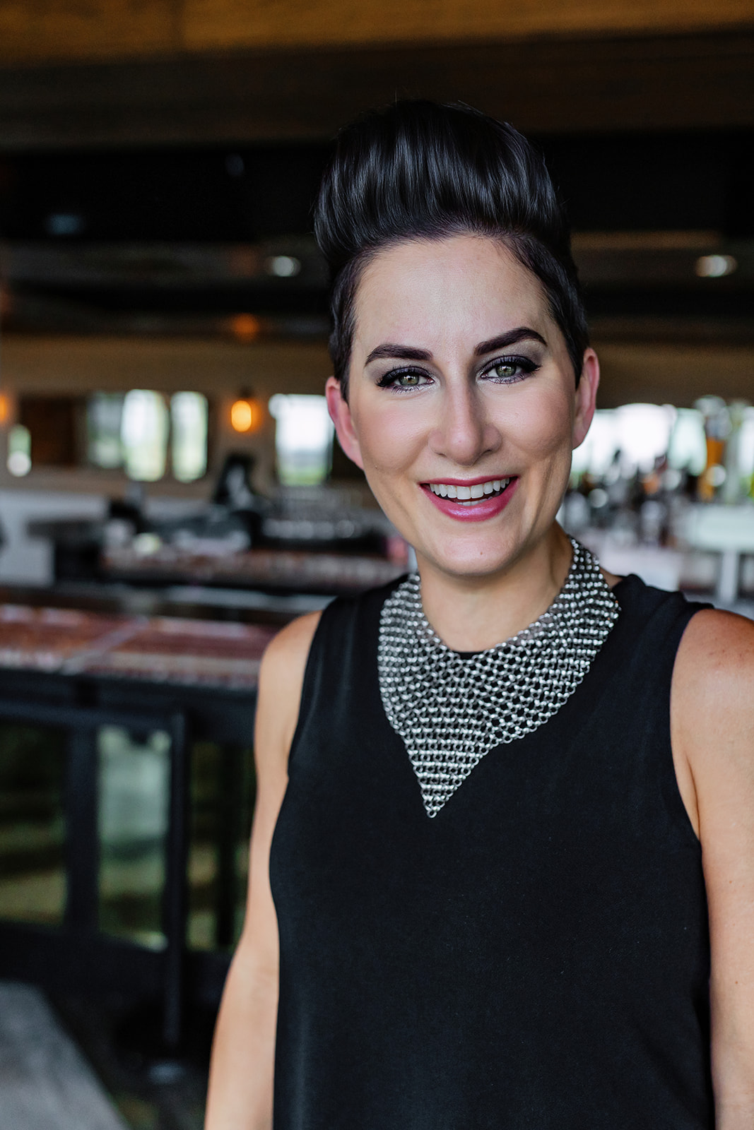 Sarah Castoral Headshot White woman with short dark hair and full makeup wearing a black tank blouse and smiling to something off-camera